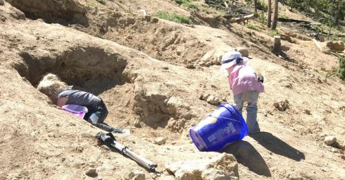 Two children digging in a sandy area, one bent over a hole and the other standing with a bucket.