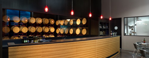 Modern wine tasting room with a wooden bar, red pendant lights, and a wall of wine barrels in the background.