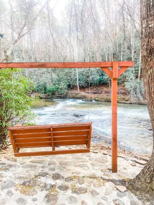 A wooden swing hangs from a frame by a river, surrounded by trees and greenery, creating a serene outdoor scene.