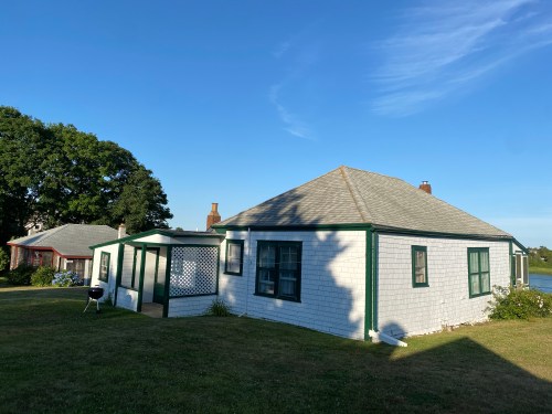 A white house with green trim, featuring a porch, surrounded by grass and trees under a clear blue sky.
