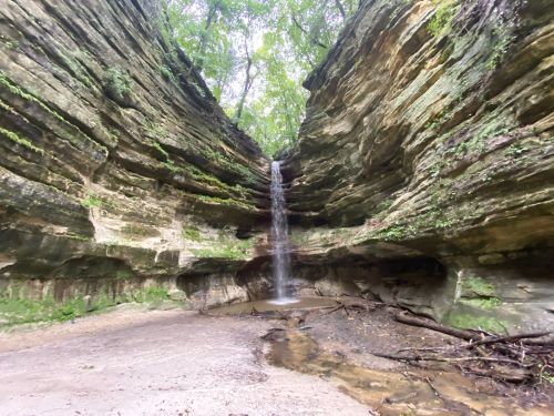 A serene waterfall cascades down rocky cliffs, surrounded by lush greenery in a tranquil canyon setting.