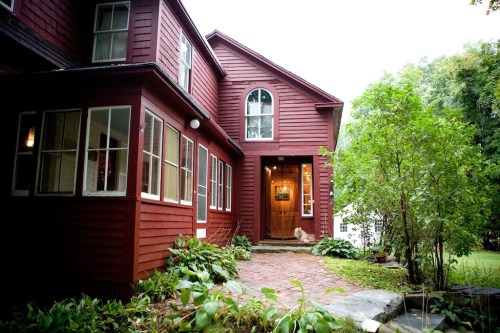 A red house with large windows, a brick pathway, and greenery leading to the entrance.