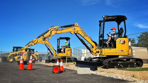 Two children operate yellow excavators on a construction site, surrounded by traffic cones under a clear blue sky.