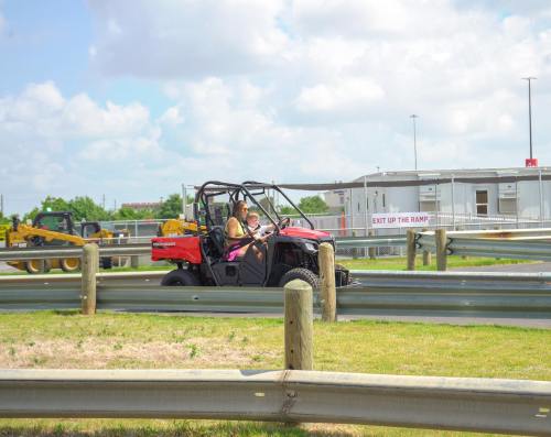 A person drives a red off-road vehicle on a track, with construction equipment and a building in the background.