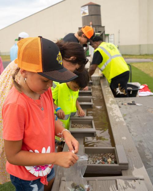 Children and adults sift through gravel at a water table, searching for treasures in a fun outdoor activity.