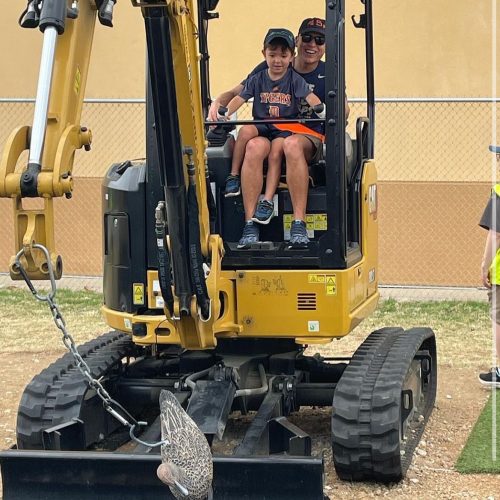 A child and an adult sit in a small excavator, smiling and enjoying their time together outdoors.