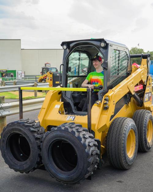 A person in a colorful shirt sits in a yellow skid steer loader at an outdoor event.