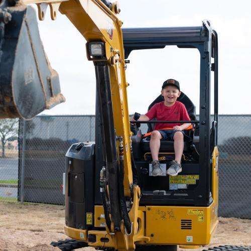 A young boy sits in a yellow excavator, smiling as he operates the machine in an outdoor setting.
