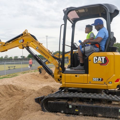 A child and an adult sit in a yellow CAT excavator, surrounded by sand at a construction site.