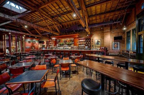 A spacious restaurant interior with wooden beams, a bar, and empty tables and chairs. Warm lighting creates a cozy atmosphere.