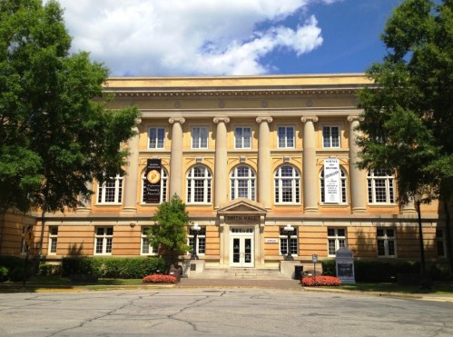 Historic building with columns, large windows, and banners, surrounded by trees and flowers under a blue sky.