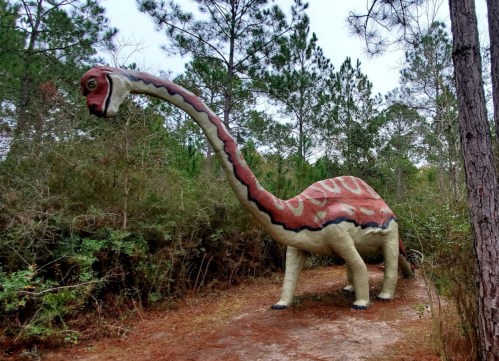 A large, colorful dinosaur statue stands among trees in a wooded area, surrounded by greenery and dirt paths.