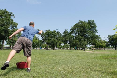A person throws a frisbee in a park, surrounded by trees and a clear blue sky.