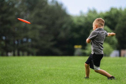 A young boy throws a frisbee in a grassy field, surrounded by trees on a cloudy day.