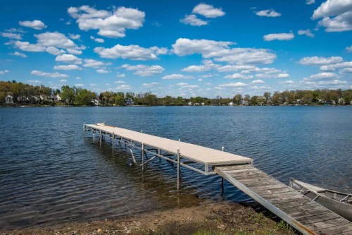 A wooden dock extends into a calm lake under a bright blue sky with fluffy white clouds.