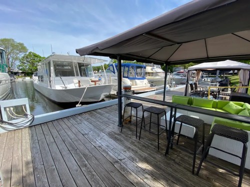 A marina scene featuring boats docked alongside a wooden deck with outdoor seating and a canopy.