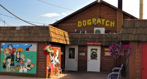 A wooden building with "DOGPATCH" sign, featuring colorful murals and hanging flower baskets at the entrance.
