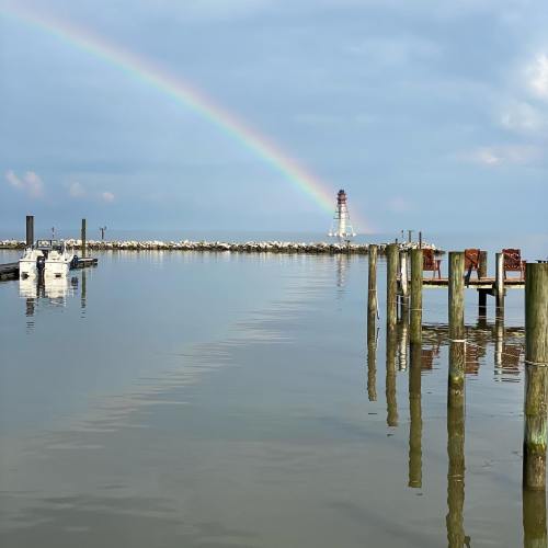 A calm waterfront scene with a rainbow arching over a lighthouse and boats docked near wooden posts.