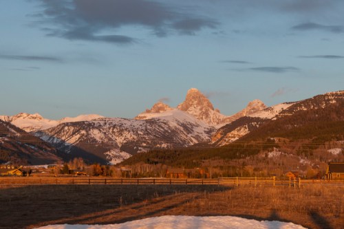 Snow-capped mountains under a golden sunset, with a grassy foreground and scattered clouds in the sky.
