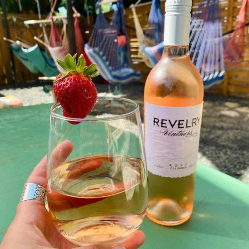 A hand holds a glass of white wine garnished with a strawberry, next to a bottle of Revelry rosé in a vibrant outdoor setting.