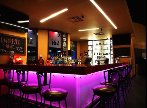 A modern bar with a glowing purple counter, high stools, and shelves of liquor bottles in a vibrant setting.