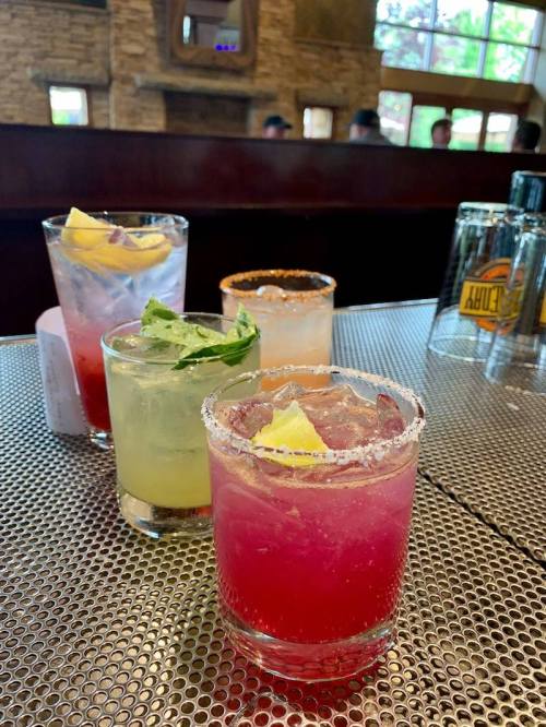 A variety of colorful cocktails with garnishes, served on a metal table in a lively bar setting.