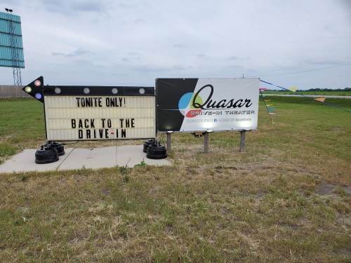 Sign for Quasar Drive-In Theater announcing "Tonite Only! Back to the Drive-In" with a grassy area and cloudy sky.