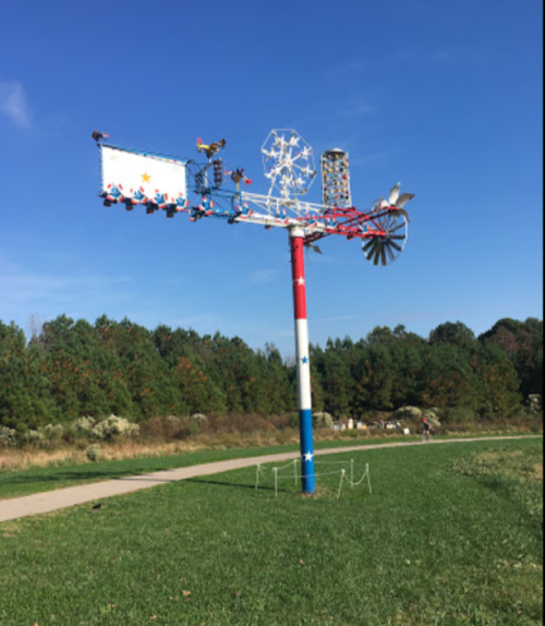 A colorful windmill sculpture with various decorative elements stands against a clear blue sky and green landscape.