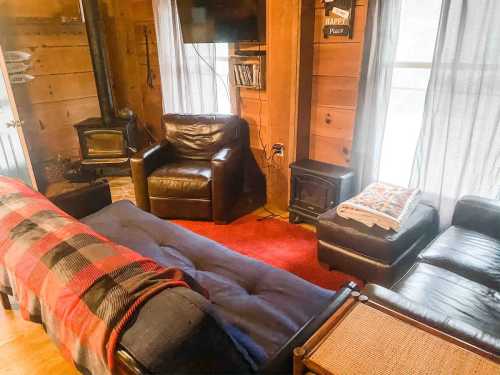 Cozy living room with leather chairs, a sofa bed, a red rug, and a wood stove, featuring wooden walls and large windows.