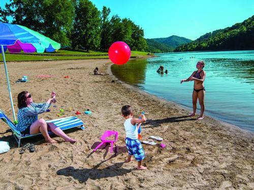 A sunny beach scene with a woman relaxing under an umbrella, a child playing with a red ball, and others enjoying the water.