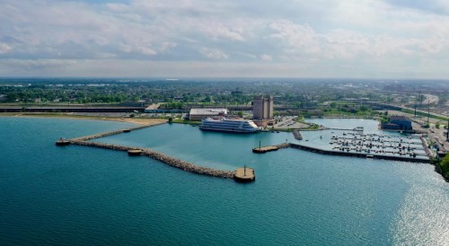 Aerial view of a harbor with a cruise ship, marinas, and cityscape in the background under a cloudy sky.