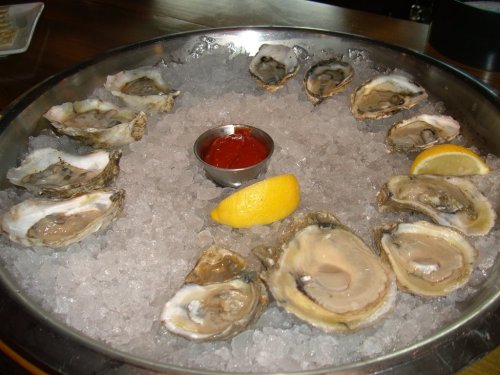 A platter of fresh oysters on ice, with a small bowl of cocktail sauce and lemon wedges.
