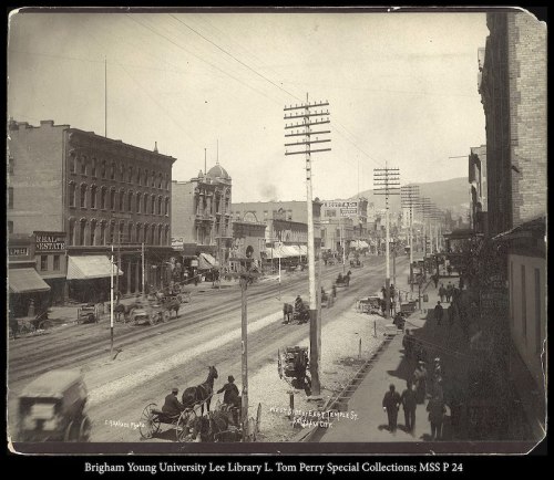 Historic street view featuring horse-drawn carriages, buildings, and power lines in a bustling town scene.
