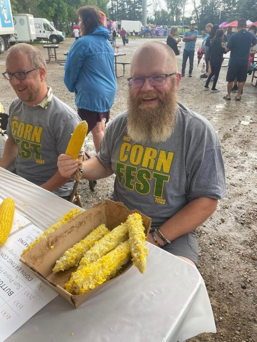 Two men at a table enjoying corn on the cob at a festival, wearing matching "Corn Fest 2022" t-shirts.