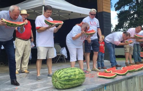 Participants compete in a watermelon-eating contest on stage, with a large watermelon in front of them.