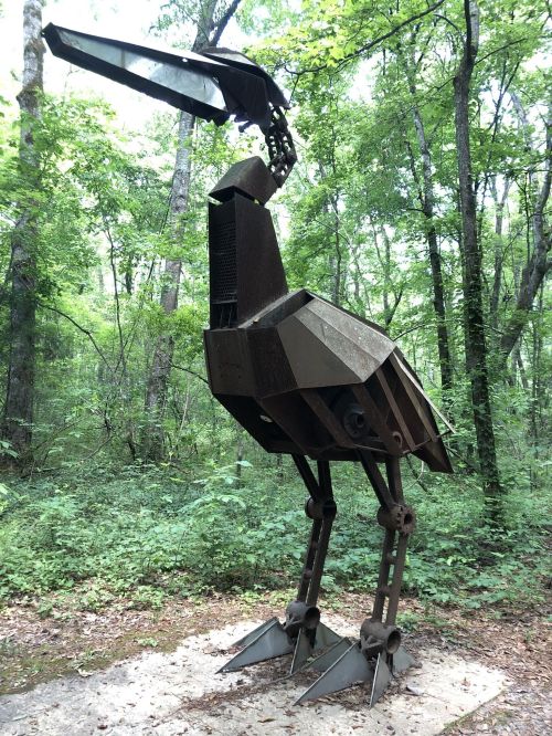 A large, metallic bird sculpture stands in a lush green forest, blending art with nature.