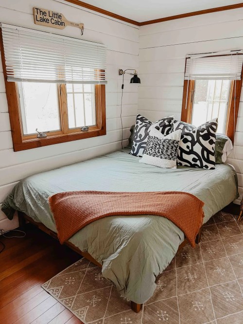Cozy bedroom in a cabin with a green bedspread, decorative pillows, and natural light from two windows.