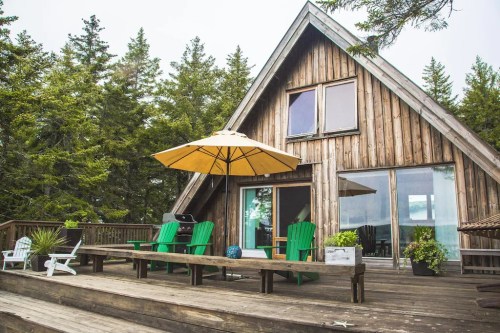 A cozy wooden A-frame cabin with green chairs and an umbrella on a deck, surrounded by tall trees.