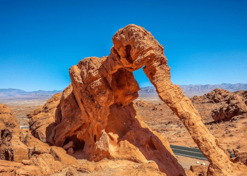 A striking red rock formation resembling an elephant trunk against a clear blue sky in a desert landscape.