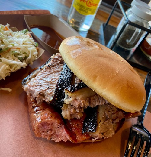 A close-up of a sandwich with layers of smoked meat on a bun, accompanied by coleslaw and barbecue sauce.