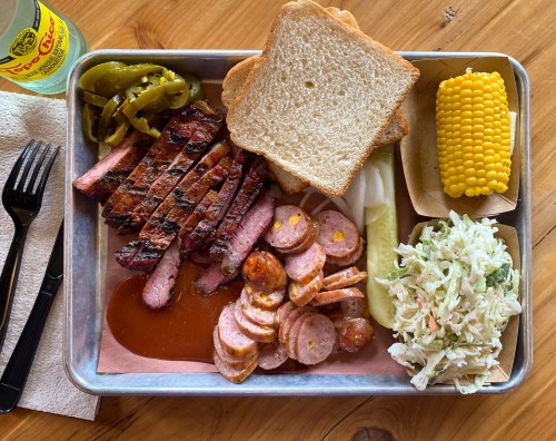 A metal tray with sliced smoked meat, sausage, pickles, jalapeños, corn, coleslaw, and slices of bread.