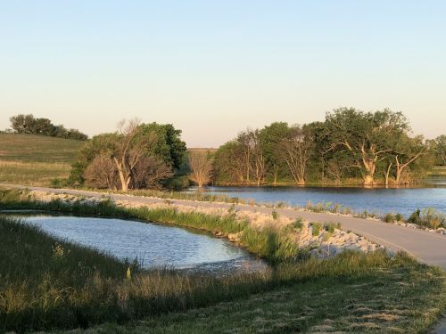 A serene landscape featuring a calm lake, grassy banks, and trees reflecting in the water under a clear sky.