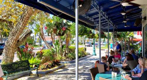 Outdoor café scene with patrons sitting at tables, surrounded by lush greenery and colorful flowers.