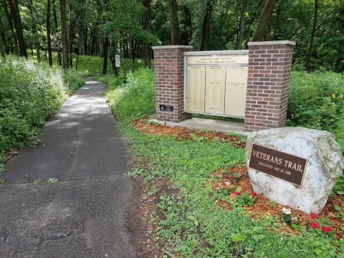 A path through a wooded area leads to a stone marker for Veterans Trail, with a plaque and informational display nearby.