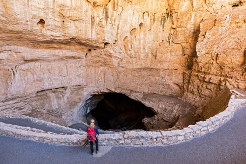 A person stands near the edge of a large cave opening, surrounded by rocky walls and a paved pathway.