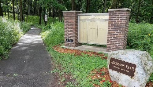 A pathway leads through greenery, with a stone sign for Veterans Trail and a brick information board nearby.