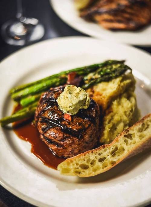 A plated meal featuring a grilled steak topped with herb butter, asparagus, mashed potatoes, and a slice of bread.
