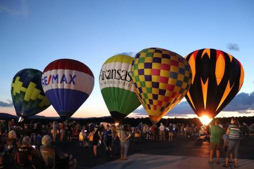 Colorful hot air balloons are illuminated at dusk, with a crowd watching in the foreground.