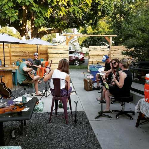 A group of musicians playing instruments outdoors in a cozy, shaded setting with trees and wooden fences.