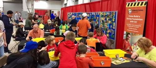 A busy event with children and adults building with colorful blocks at tables, surrounded by a vibrant backdrop and banners.